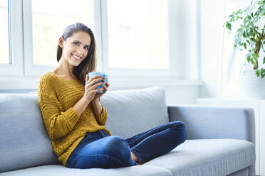 Beautiful young woman sitting on sofa drinking coffee and looking at camera - BSZF01532