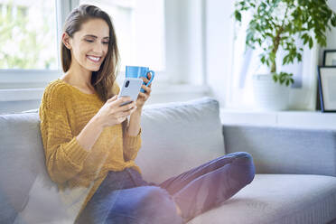 Smiling young woman sitting on sofa with smartphone and cup of coffee - BSZF01535