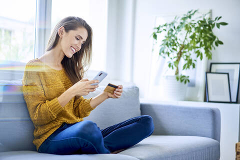 Schöne glückliche Frau, die mit Smartphone und Kreditkarte online bezahlt, während sie auf dem Sofa sitzt, lizenzfreies Stockfoto