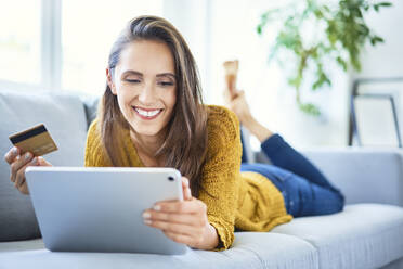 Fröhliche junge Frau benutzt Kreditkarte und Tablet, um von zu Hause aus online einzukaufen - BSZF01537
