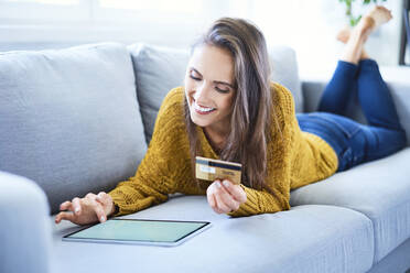 Lächelnde junge Frau, die auf dem Sofa liegend mit Kreditkarte und Tablet online einkauft - BSZF01540