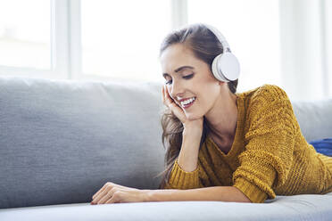 Cheerful young woman lying on sofa and listening to music with headphones - BSZF01543