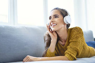 Happy young woman listening to music with headphones while lying on sofa - BSZF01544