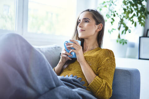 Schöne junge Frau, die eine Tasse Kaffee genießt, während sie auf dem Sofa unter einer Decke liegt, lizenzfreies Stockfoto