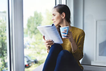 Young woman looking out the window while reading book at home - BSZF01548