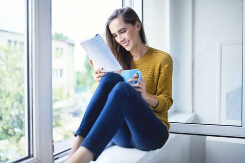 Lächelnde junge Frau sitzt auf der Fensterbank, liest ein Buch und trinkt Kaffee - BSZF01549