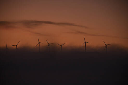 Spain, Province of Cadiz, Tarifa, Silhouettes of wind turbines standing against moody sky at foggy dawn - KBF00613