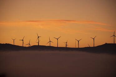 Spain, Province of Cadiz, Tarifa, Silhouettes of wind turbines standing against moody sky at foggy dawn - KBF00614