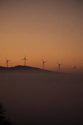 Spain, Province of Cadiz, Tarifa, Silhouettes of wind turbines standing against moody sky at foggy dawn - KBF00615