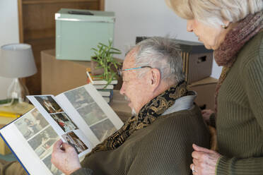 Senior couple looking at photo album surrounded by cardboard boxes in an empty room - MAMF00815
