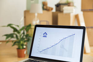Rising line graph on laptop screen in front of cardboard boxes in a new home - MAMF00846