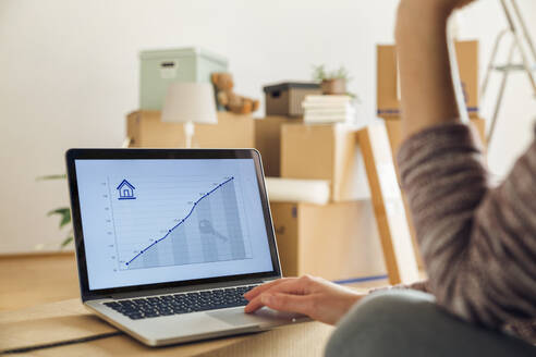 Frau mit Laptop und ansteigendem Liniendiagramm auf dem Bildschirm in einer neuen Wohnung - MAMF00849