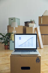Rising line graph on laptop screen in front of cardboard boxes in an empty room in a new home - MAMF00853