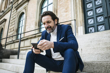 Porträt eines bärtigen reifen Geschäftsmannes, der auf einer Treppe sitzt und auf sein Handy schaut - JLOF00323