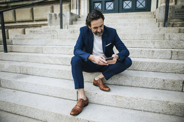 Bärtiger reifer Geschäftsmann im blauen Anzug sitzt auf einer Treppe und schaut auf sein Handy - JLOF00338