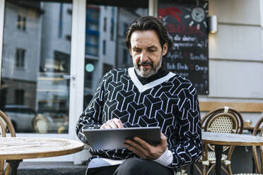 Porträt eines modischen reifen Mannes, der ein digitales Tablet in einem Straßencafé benutzt - JLOF00344
