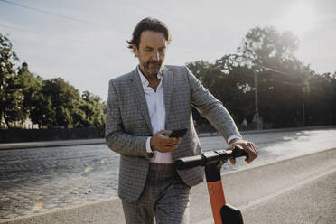 Geschäftsmann schaut auf sein Smartphone und hält einen E-Scooter in der Stadt - JLOF00372