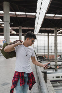 Young man waiting at train station, using smartphone - JMHMF00009