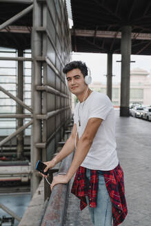 Young man waiting at train station with smartphone and wearing headphones - JMHMF00011