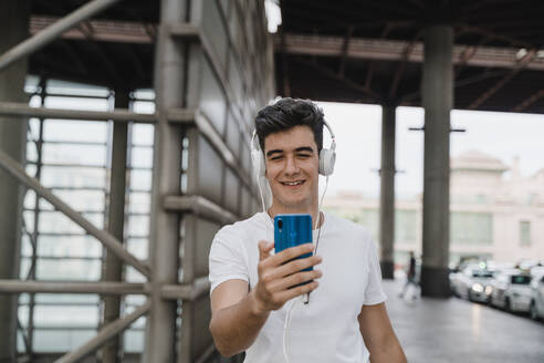 Young man waiting at train station with smartphone and wearing headphones - JMHMF00012