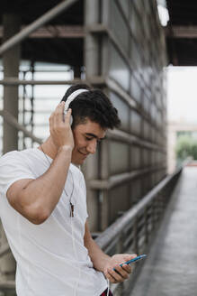 Young man waiting at train station with smartphone and wearing headphones - JMHMF00013