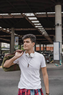 Young man with a bag at train station wearing headphones - JMHMF00014