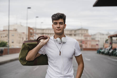 Young man with a bag at train station wearing headphones - JMHMF00016