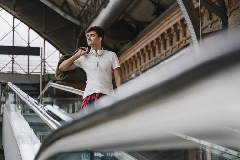 Young man standing on escalator at train station and wearing headphones - JMHMF00018