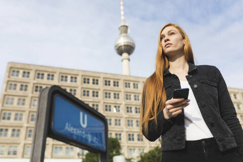 Porträt einer rothaarigen jungen Frau mit Smartphone am Alexanderplatz, Berlin, Deutschland, lizenzfreies Stockfoto
