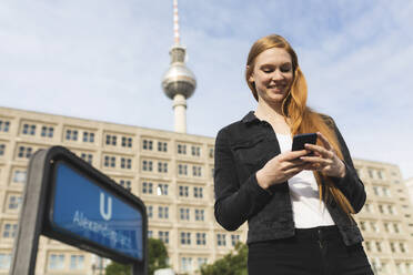 Porträt einer lächelnden jungen Frau am Alexanderplatz, die auf ihr Handy schaut, Berlin, Deutschland - WPEF01998