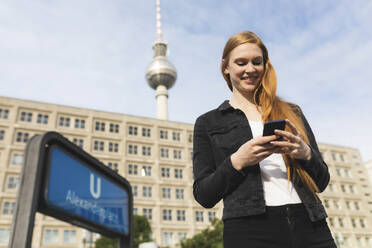 Portrait of smiling young woman at Alexanderplatz looking at cell phone, Berlin, Germany - WPEF01998