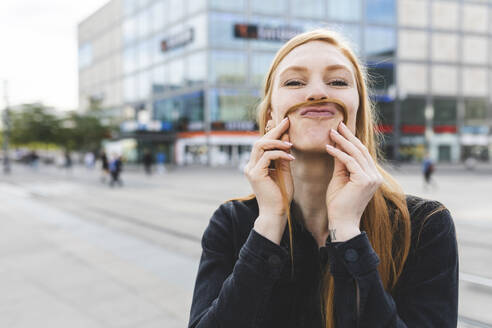 Porträt einer rothaarigen jungen Frau, die sich mit ihren Haaren einen Schnurrbart macht, Berlin, Deutschland - WPEF02000