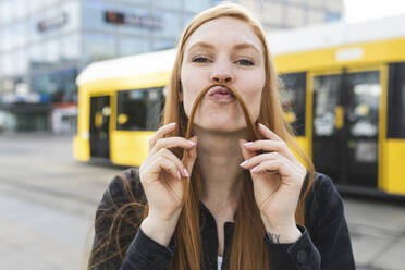 Portrait of redheaded young woman making mustache with her hair, Berlin, Germany - WPEF02001