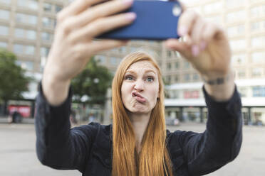 Porträt einer rothaarigen jungen Frau, die einen Schmollmund zieht, während sie ein Selfie mit ihrem Smartphone macht, Berlin, Deutschland - WPEF02004