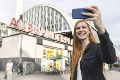 Porträt einer rothaarigen jungen Frau, die ein Selfie mit ihrem Smartphone am Alexanderplatz macht, Berlin, Deutschland, lizenzfreies Stockfoto
