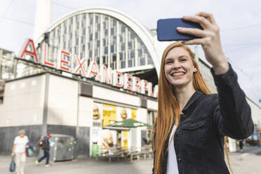 Porträt einer rothaarigen jungen Frau, die ein Selfie mit ihrem Smartphone am Alexanderplatz macht, Berlin, Deutschland - WPEF02009