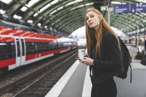Porträt einer rothaarigen jungen Frau mit Smartphone und Kaffee zum Mitnehmen, die am Bahnsteig wartet, Berlin, Deutschland - WPEF02010