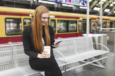 Rothaarige junge Frau mit Kaffee zum Mitnehmen wartet am Bahnsteig und benutzt ihr Smartphone, Berlin, Deutschland - WPEF02013