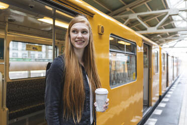 Porträt einer rothaarigen jungen Frau mit Kaffee zum Mitnehmen beim Aussteigen aus dem Zug, Berlin, Deutschland - WPEF02014