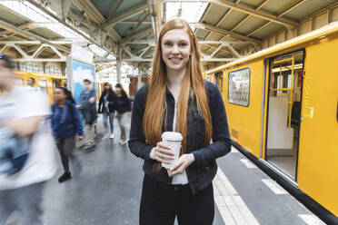 Porträt einer rothaarigen jungen Frau mit Kaffee zum Mitnehmen auf dem Bahnsteig, Berlin, Deutschland - WPEF02015