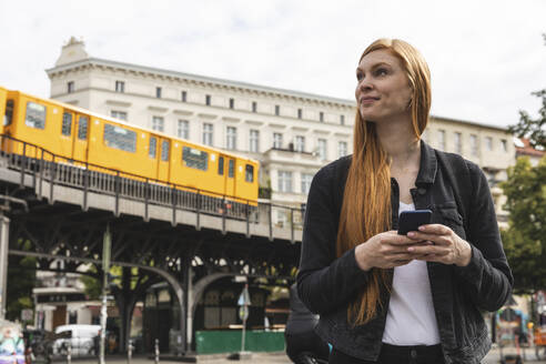 Porträt einer rothaarigen jungen Frau mit Smartphone in der Stadt, Berlin, Deutschland - WPEF02016