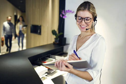 Smiling receptionist talking with headset while taking notes - JSRF00671