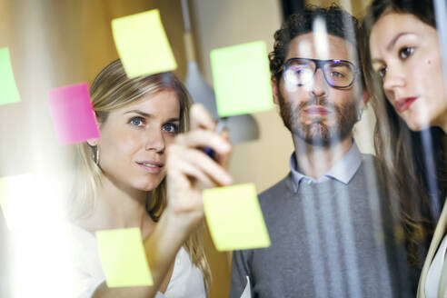 Three business people brainstorming together with sticky notes on a glass wall - JSRF00687