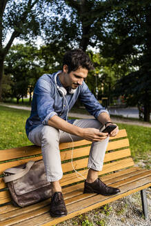 Lächelnder Mann sitzt auf einer Parkbank und benutzt sein Smartphone - GIOF07172