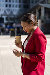Lächelnde Geschäftsfrau mit Kaffee zum Mitnehmen, die auf ihr Mobiltelefon schaut, London, UK - MAUF02941