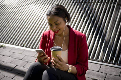 Lächelnde Geschäftsfrau mit Kaffee zum Mitnehmen, die ein Mobiltelefon und Ohrstöpsel benutzt, London, UK, lizenzfreies Stockfoto