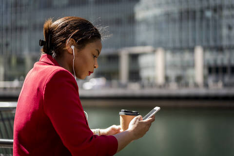 Profile of businesswoman with coffee to go using mobile phone and earbuds, London, UK stock photo
