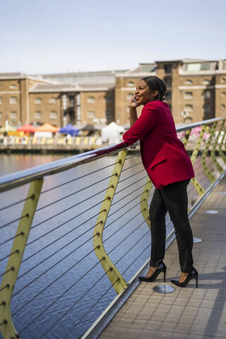 Lächelnde Geschäftsfrau am Telefon auf einer Brücke stehend und nach oben schauend, London, UK, lizenzfreies Stockfoto