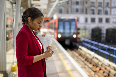 Geschäftsfrau steht auf einem Bahnsteig und schaut auf ein Mobiltelefon, London, UK - MAUF02964