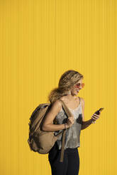 Lachende junge Frau mit Rucksack schaut auf Handy vor gelbem Hintergrund - DAMF00143